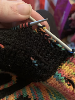 The first cast off stitches of the round worked using both the rainbow yarn and the black yarn. It's situated over a black stockinette background section of the bag.