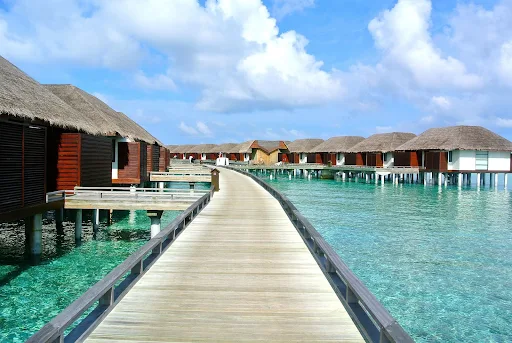 A serene view of the Maldives, featuring luxurious overwater bungalows built on stilts above crystal-clear turquoise waters. A wooden walkway leads through the resort, surrounded by the calm ocean. The sky is bright blue with fluffy white clouds, creating a picturesque tropical paradise setting.