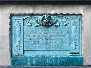 A photo of a blue plaque commemorating the original St Paul’s Cross.  Above the text is the face of an angelic looking figure.  Photo by Kevin Nosferatu for the Skulferatu Project.