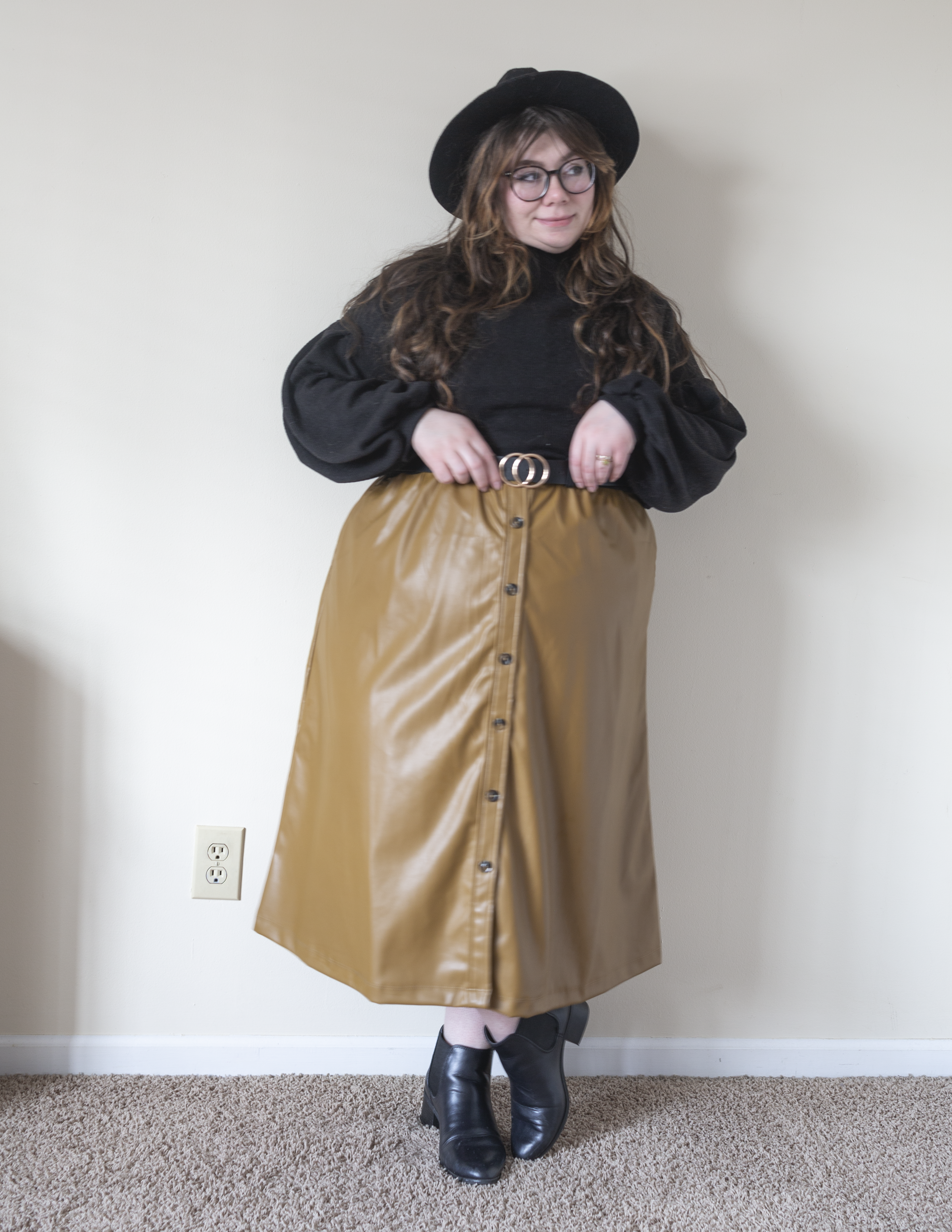 An outfit consisting of a black balloon sleeve sweater over a camel brown short sleeve faux leather -line dress worn as a skirt, with black sheer tights and black Chelsea boots.