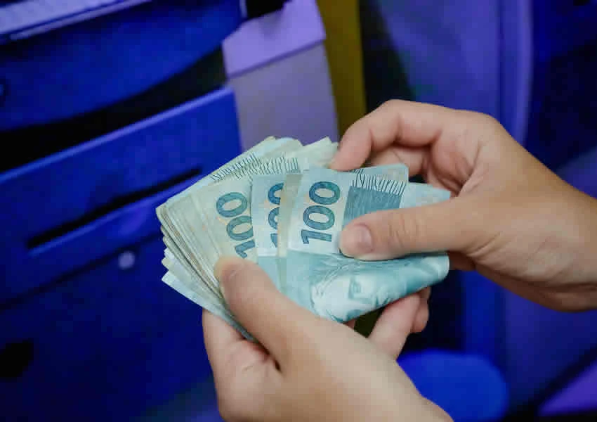 Imagem mostra duas mãos contando notas de 100 reais em frente a um Caixa eletrônico.