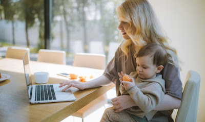 A mom works at her laptop while holding her child who is eating an orange.