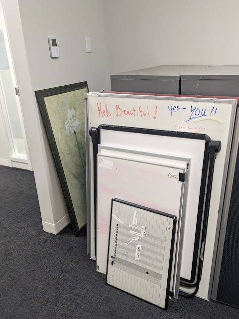 Picture of office hall. Five used whiteboards are leaning against filing cabinets. The top edge of the larger whiteboard has written on it with red marker: "Hey Beautiful" and with blue marker, "Yes, you!!"