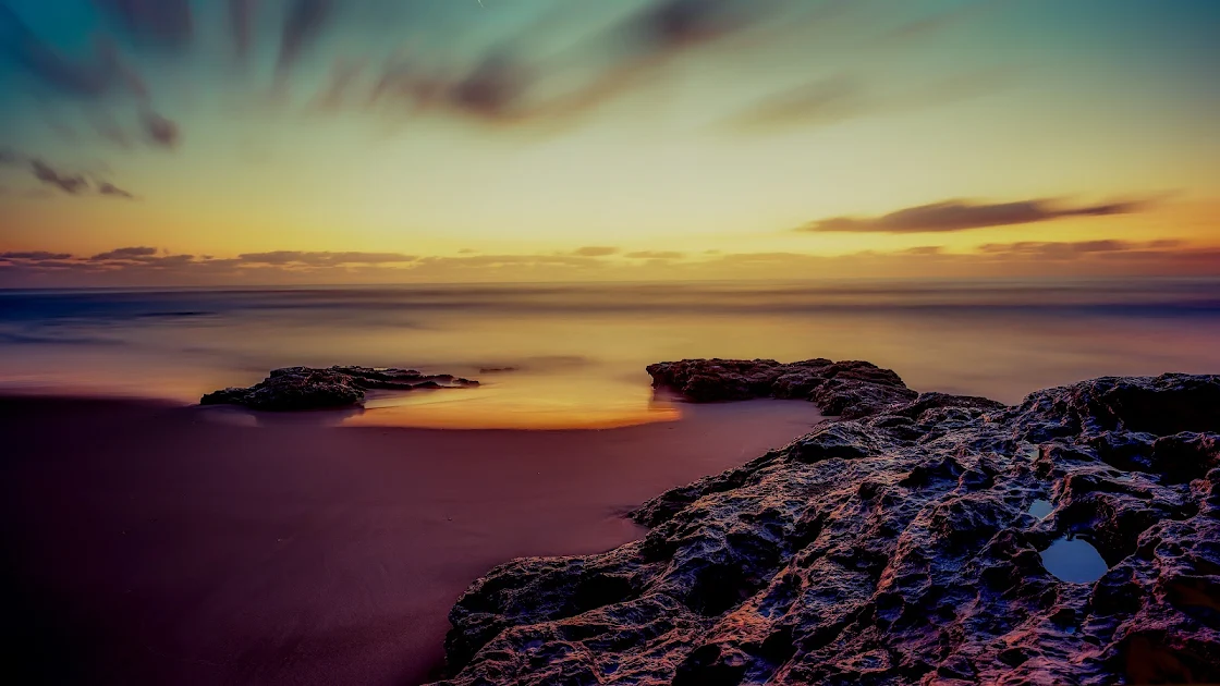 Golden and blue hues of dusk sky reflecting over serene coastal waters, bordered by rugged rocks in high-definition 4K.