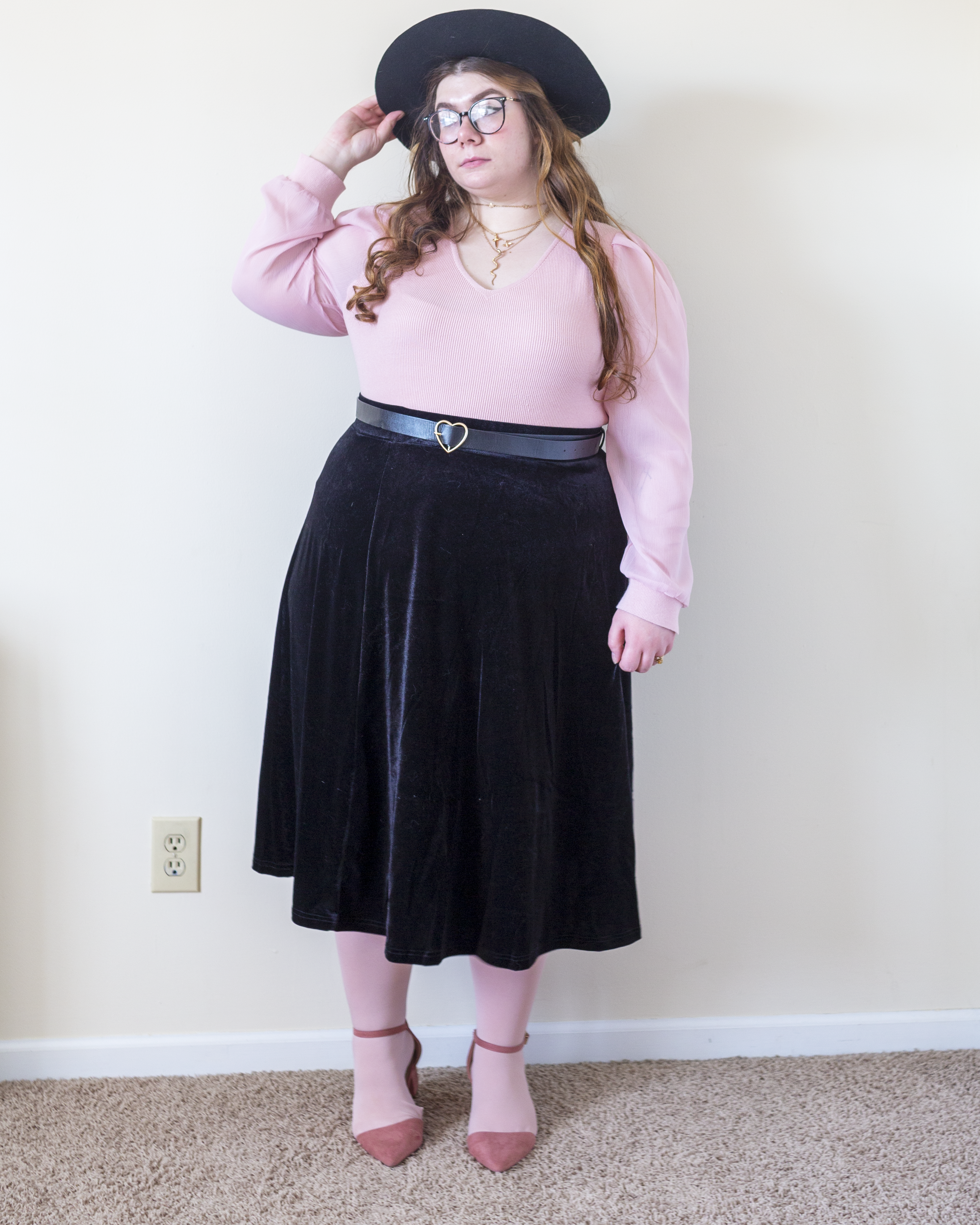 An outfit consisting of a pastel pink v-neck sweater with sheer sleeves, tucked into a black velvet skater midi skirt and pastel pink ankle strap heels.