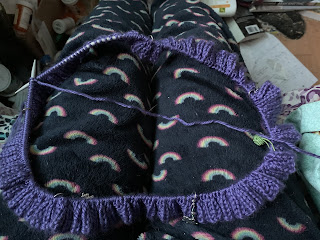 A long circular knitting needle with several rounds of knitting in a smooth purple yarn done. There are several stitch markers in place around the needle, 4 silver, 1 green "yarn ball" and a darker green one.