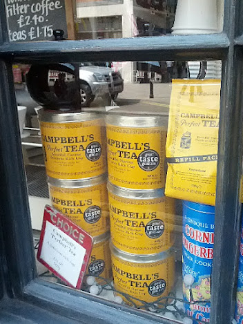 Items in a storefront windows: tins of Campbell's tea, a chalkboard with the price of takeout coffee and tea, a large tea of Cornish gingerbread.