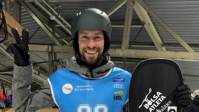 André posa para foto segurando prancha de snowboard e fazendo um sinal de número 2 com a mão direita. Ele veste um casaco cinza com um colete azul por cima e um capacete preto