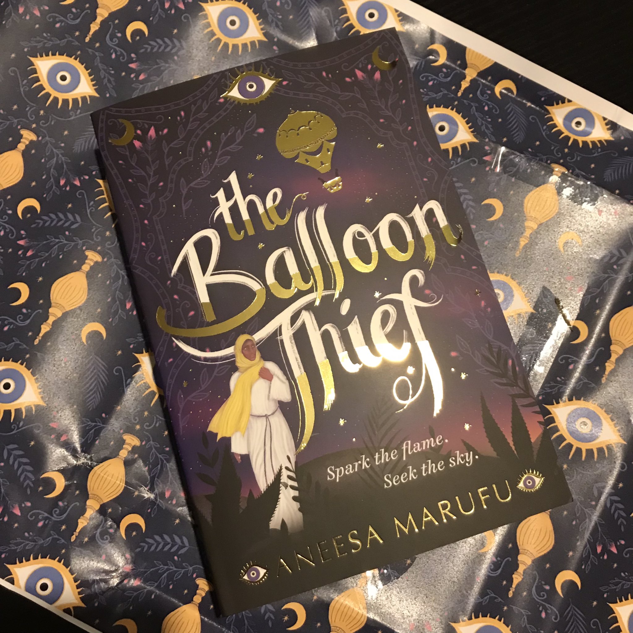 The Balloon Thief by Aneesa Marufu laying on flattened navy blue wrapping paper patterned with yellow and blue eyes and stylised urn with crescent moons repeated, on a black wooden table. The book is diagonal, top left to bottom right.