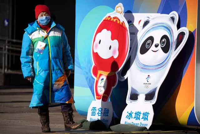 Mulher chinesa vestida de azul parada em frente aos mascotes dos Jogos Olímpicos e Paralimpicos