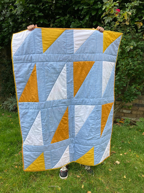 A blue, yello and white triangle quilt is being held up by a child.  They are standing on grass, with a brick wall behind.  You can only see the child's feet poking out under the quilt.