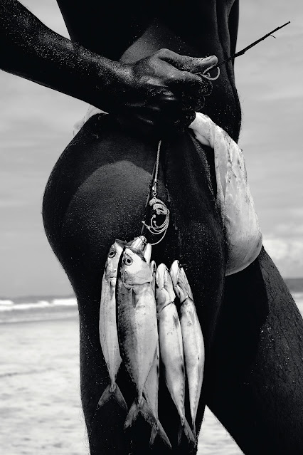 Homem saindo do mar, com uma sunga que deixa os pelos de sua virilha aparente. Ele segura um anzol do lado do corpo com peixes pendurados.