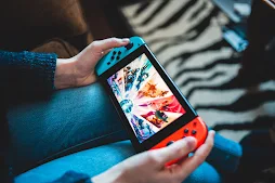 Someone wearing blue jeans playing using the tablet-like Nintendo Switch. The switch has detachable controls and the left controller is blue and the right one is red.