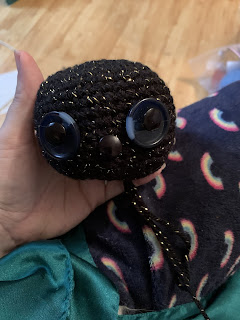 What appears to be a crocheted ball with buttons sewn into positions resembling two eyes and a nose. The black yarn has a gold metallic strand through it