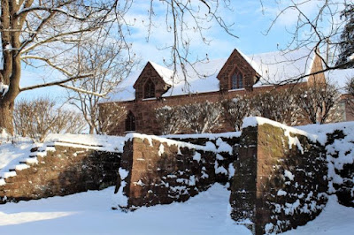 Stone wall with shorter walls at right angles. Gothic style stone building is on the other side of wall. Snow covers the ground and tops of the walls and building.