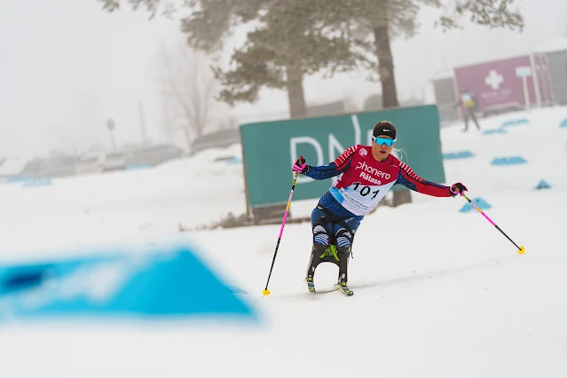 Oksana Masters esquia em pista de neve. Ela usa esquis adaptado com uma especie de cadeira e está usando um bastão para se equilibrar em uma curva. Maters veste uma malha vermelha e azul, que lembra a bandeira dos Estados Unidos, com um colete com o número 101 por cima