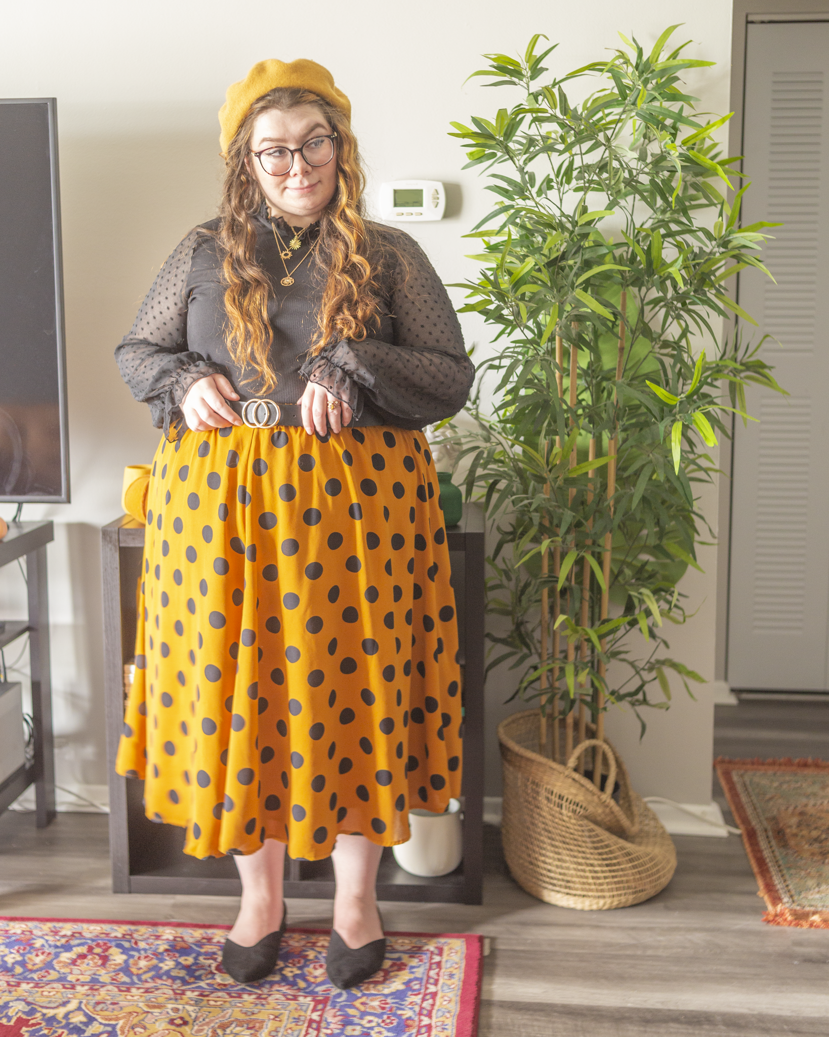 An outfit consisting of a muted yellow wool beret, a black sheer bishop sleeve with polka dots high beck blouse tucked into a black on cinnamon orange midi skirt and black sling back pointed toe flats.