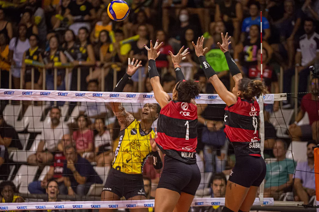 Martinez ataca bola contra um bloqueio duplo do flamengo. A jogadora do praia usa uma camisa amarela e preta e as flamenguistas usam blusa listrada vermelha e preta