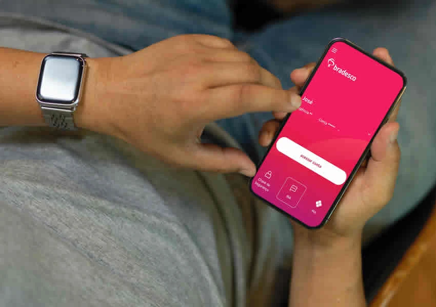 Imagem mostra um homem sentado segurando um smartphone com uma das mãos e com e outra o manuseando com o app do bradesco aberto.