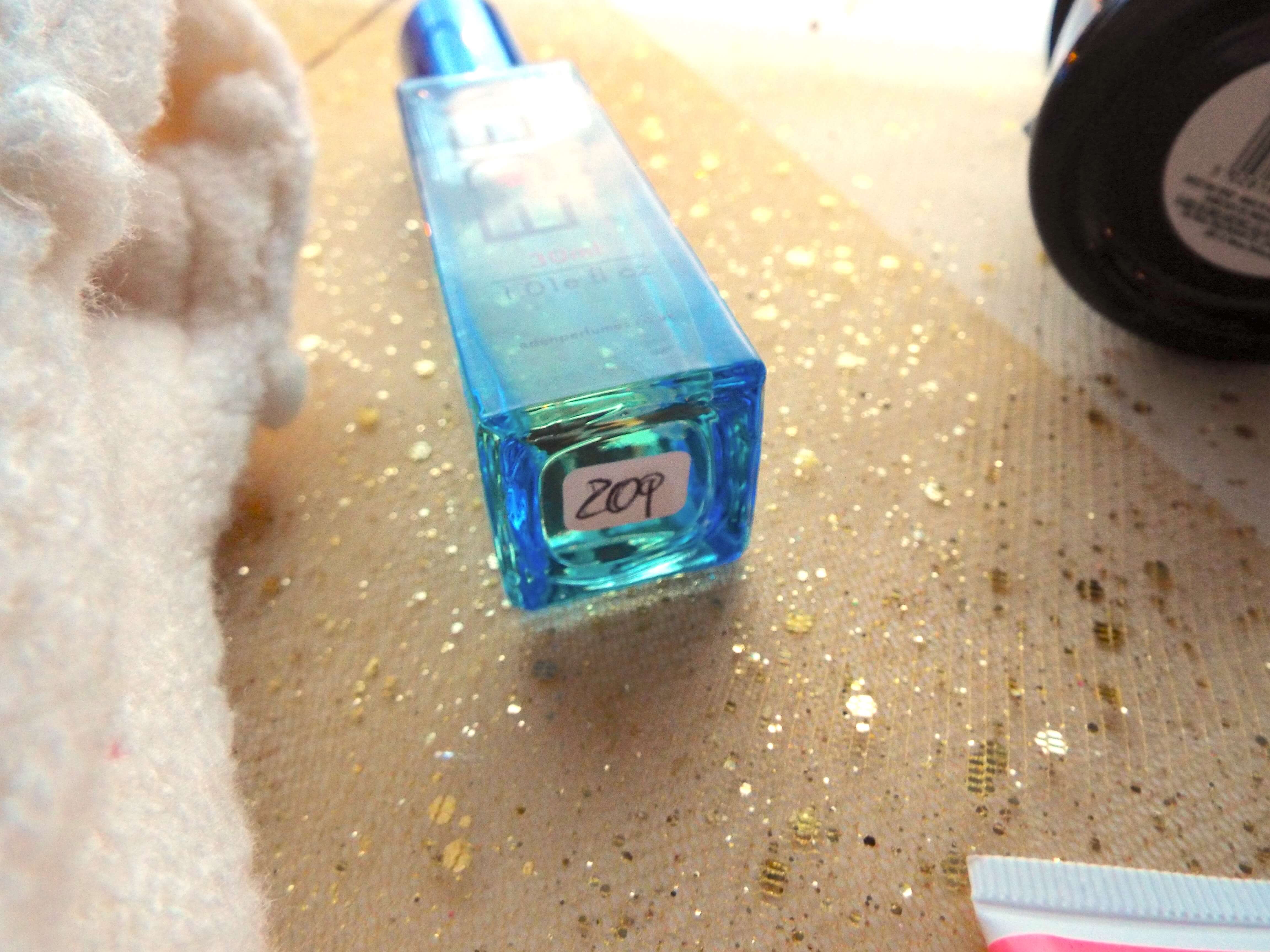 Eden vegan elixir perfume in 209, in blue bottle, lay on gold sparkly table runner, adjacent to cream cardigan.