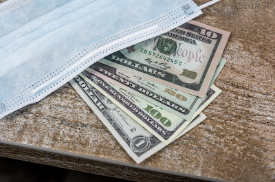 Dollar bills on a brown table top partially covered by a surgical face mask.