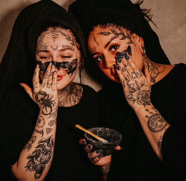 Hollie and Lorna, founders of Wild and Wood Skincare, applying a black face mask. They both have lots of amazing black tattoos over their arms and faces.