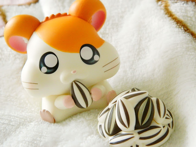 A photo showing an orange and white hamster character called Hamtaro. He has chubby cheek pouches and his holding a sunflower seed. There is a pile of sunflower seeds in front of him.