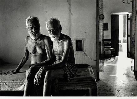 Dois homens sentados na cama de um quarto, encarando fixamente a câmera. Ao lado direto deles há uma porta que mostra parte dos outros cômodos da casa.