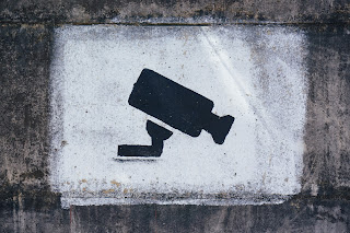 A black security camera stencilled onto a painted white cement background that is rectangular in shape. Around the white rectangle is more cement that is more dirty and grimy.