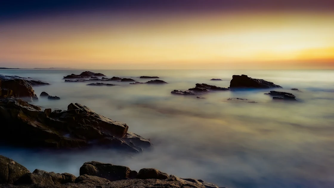 A breathtaking 4K image of a serene sunset over a misty seascape with rocky shores.