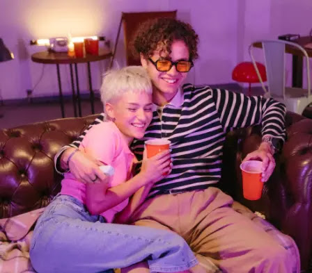 Pareja de hombre y mujer adolescentes sentados en un sillón de piel color café, sosteniendo cada uno un vaso desechable de refresco en la mano