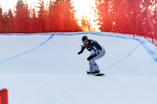 Huckaby encima de sua prancha descendo uma pista de snowboard cross. o trecho da pista é uma reta após uma curva, sinalizados com uma tinta azul