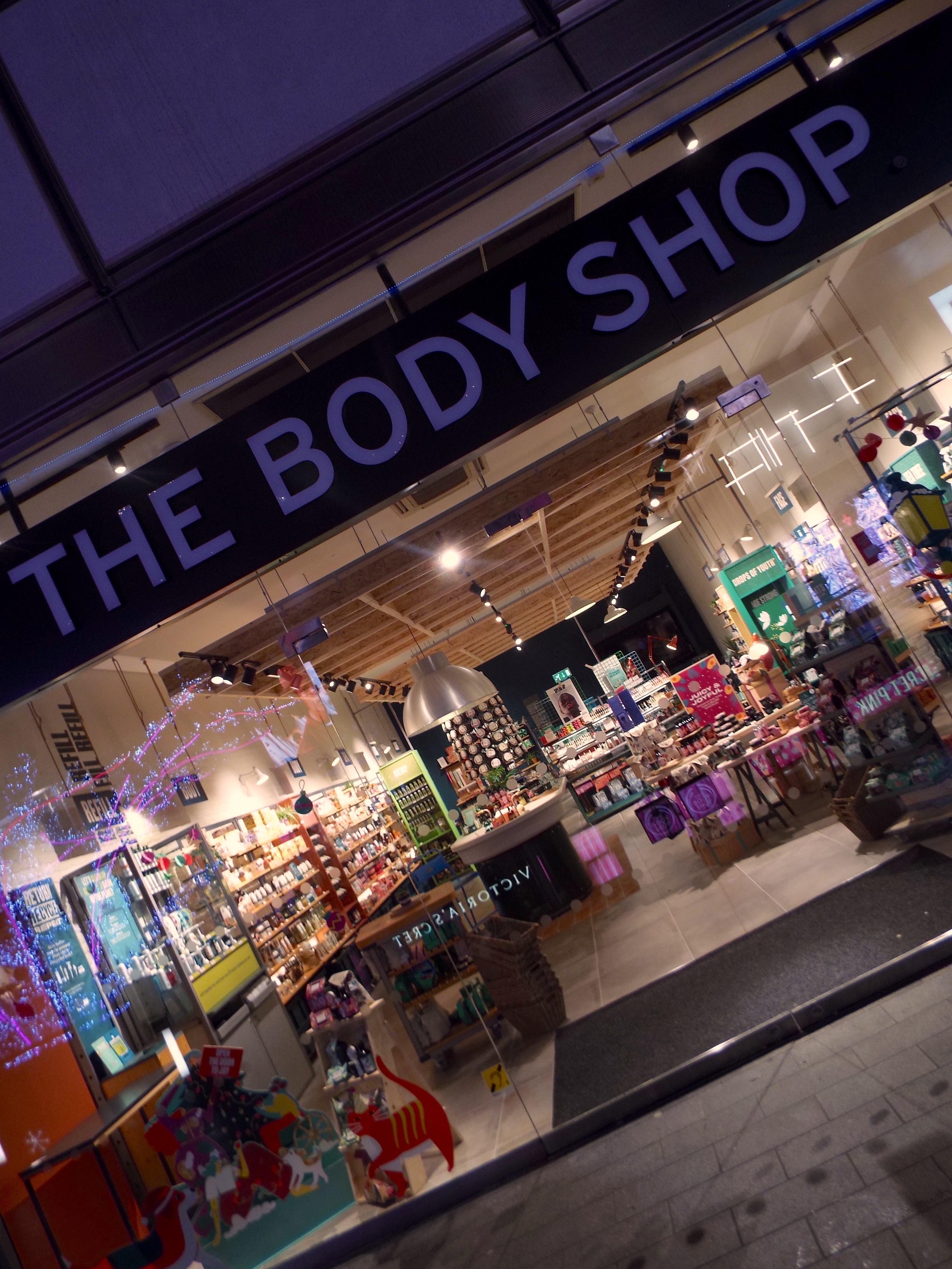 The Body Shop Liverpool One lit up on a dark Christmas evening with the surrounding Christmas lights reflecting in the window and colourful displays inside.