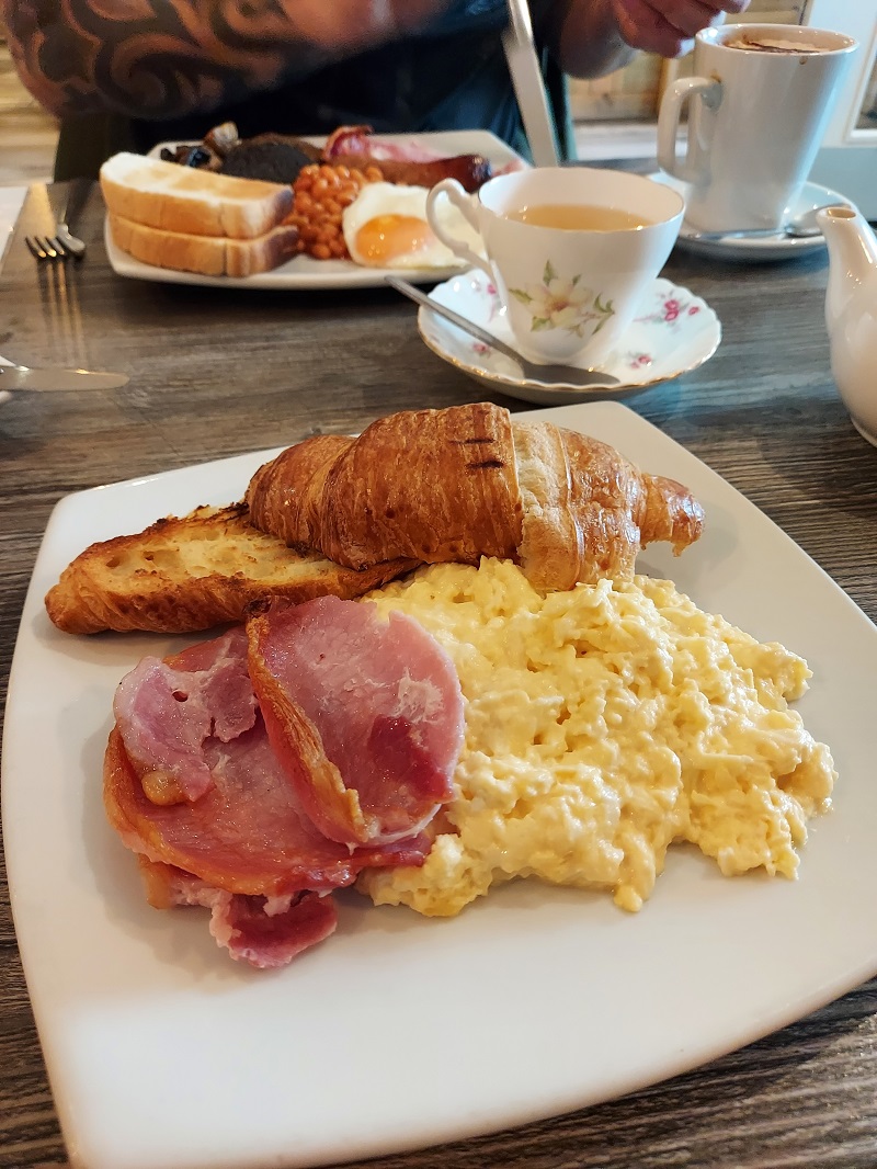 A white plate with a croissant, bacon and scrambled eggs on it. In the background is a Full Scottish breakfast at the Pier in Aberdeen