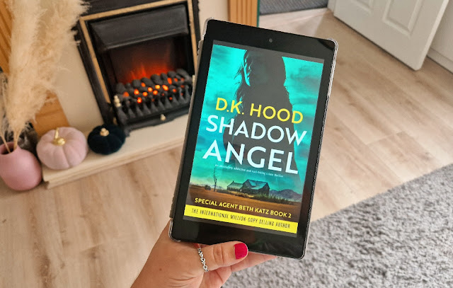 Beth is holding a kindle with the cover for shadow angel by D.K Hood on the screen. The cover shows the silhouette of a woman in a green sky over a cabin