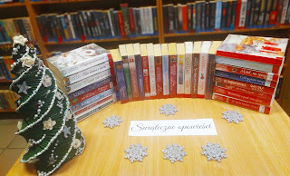 Trzy stosy książek o tematyce Świat Bożego Narodzenia leżą na stoliku. Po lewej choinka. Tło: regały z książkami. Przed książkami napis Świąteczne opowieści.