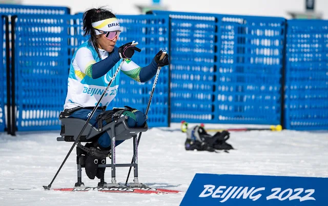 Aline esquia usando esquis adaptados com uma espécie de cadeira. Ela usa um agasalho azul e branco e um colete com o número 25