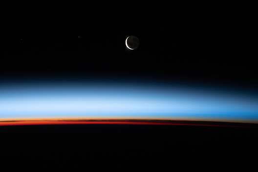 Shot from the International Space Station flying over New Zealand, a crescent Moon shines hard against the darkness of space. Below, Earth's atmosphere, in twilight, splits into a palette of sunset hues.