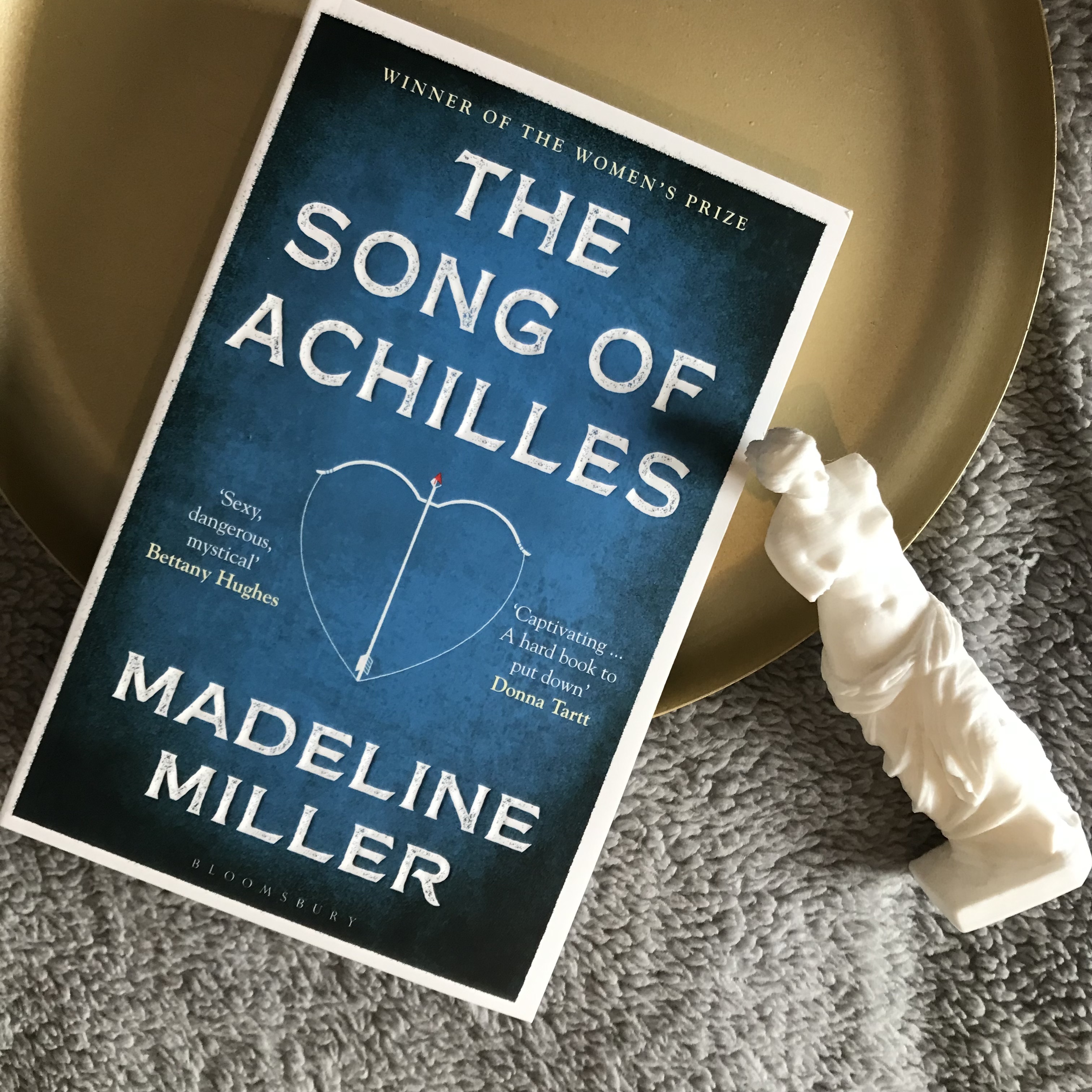 A photo of The Song of Achilles by Madeline Miller, half on a gold, circular tray, and half on a fluffy, light grey duvet. The book is diagonal, with the top facing the top right corner of the photo, and the bottom facing the bottom left corner of the photo. Next to the book, on the right, is a small, white Venus de Milo staue.