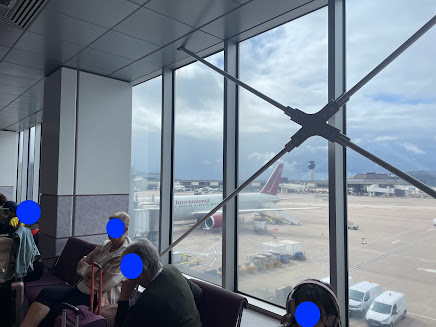 An airport departure lounge. Out of the window, a plane can be seen. People are sitting alongside the window on benches. lots of bags. (All faces have been obscured.)