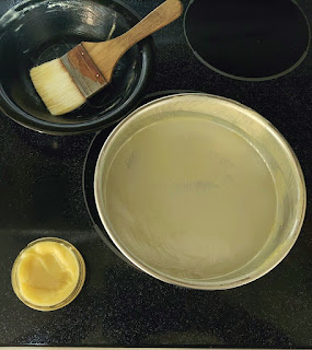 A pan smeared with the cake goop, jar of cake goop and black bowl with the used pastry brush sitting in it.