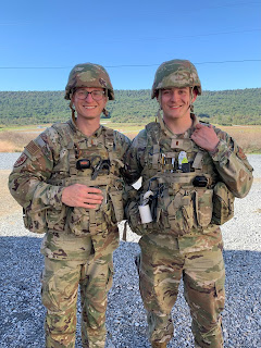 Two individuals in camouflage military uniforms, with helmets, are standing side by side and smiling at the camera. They appear to be soldiers in a training or operational setting, with a landscape of trees and a clear sky in the background.