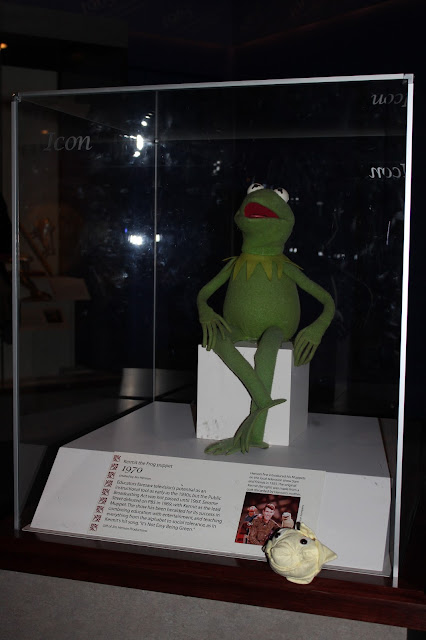 Little Star (a smiling yellow star) sits in front of a vitrine holding Kermit the Frog.  Kermit is green and looks much like a real frog except he has more human like hands/fingers.  He sits with his legs crossed and his mouth is red and pink and his eyes are half ping pong balls with black cloud like pupils.  There is a small text panel talking about Kermit on his exhibit mount base.