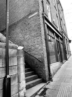 A black and white photo showing a street with a wall in the foreground, a set of steps going up and beyond that some brick buildings.  There is a sign on the wall of the building by the steps that reads Dog Leap Stairs.  Photograph by Kevin Nosferatu for the Skulferatu Project.