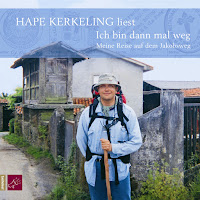 Hape Kerkeling, der mit Hut und Wanderstock ausgestattet in einer grünen Landschaft steht und in die Kamera lächelt.