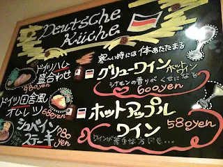 Chalkboard style menu with Japanese text in red, white, and red listing the names of different foods and their prices. Some drawings of food are shown next to the menu items. There is a German flag at the top of the chalkboard menu.