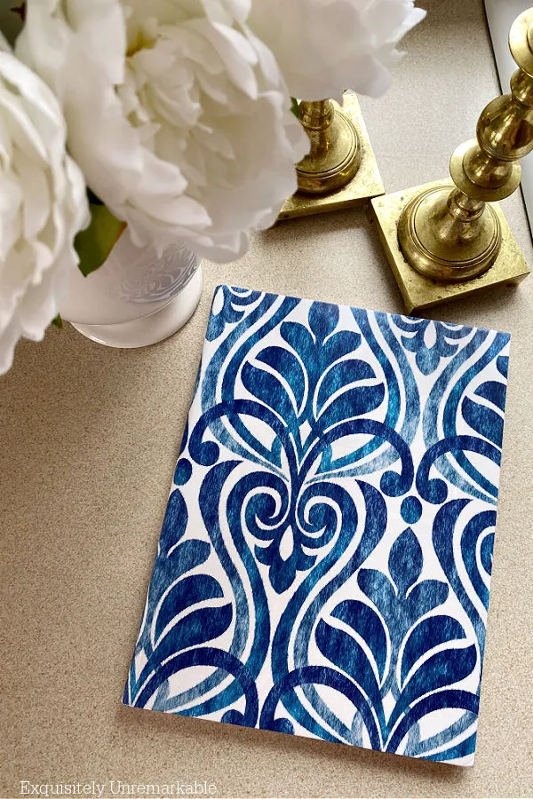 Paper covered notebook on counter between gold candlesticks and flower filled vase