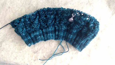 A fingering-weight lace hat in progress on circular needles, knit in a deep green yarn.