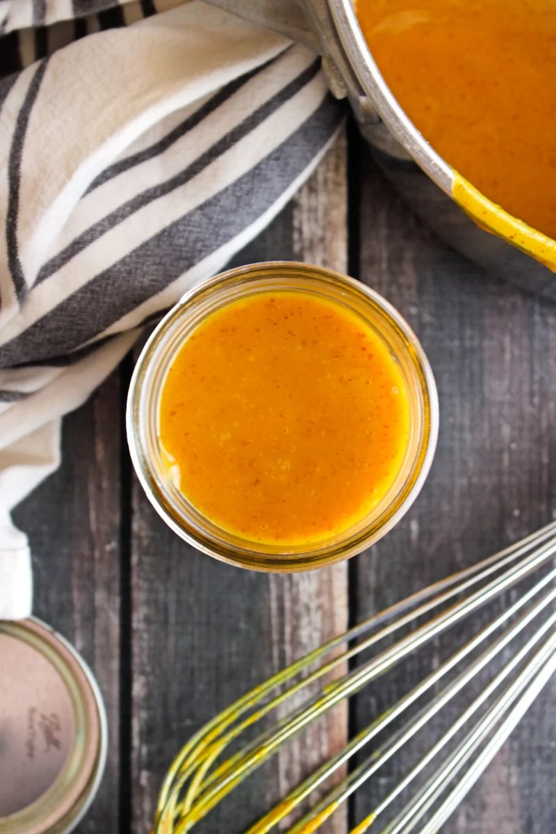 Honey mustard barbecue sauce in a small mason jar on a wood background with a blue striped kitchen towel.