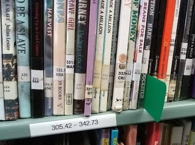 Library books on shelf, organized according to Dewey Decimal classifications that are displayed via tags on the books' spines. A green-plastic browser or 'shelf-marker' juts out from between two books, and a label on the shelf-edge identifies the span of Dewey Decimal numbers to be found among books on that shelf: '305.42-342.73.'